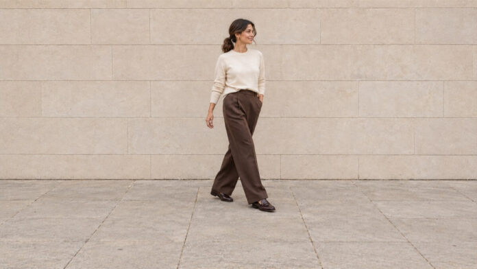 Woman with a low ponytail walking in an ivory jumper, chocolate wide-leg trousers and brown loafers.