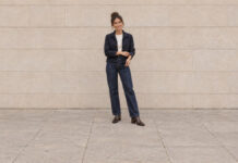 Woman with her hair in a high bun adjusting a navy jacket cuff, wearing straight-leg jeans and brown ankle boots.