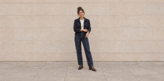 Woman with her hair in a high bun adjusting a navy jacket cuff, wearing straight-leg jeans and brown ankle boots.
