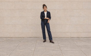Woman with her hair in a high bun adjusting a navy jacket cuff, wearing straight-leg jeans and brown ankle boots.