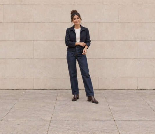 Woman with her hair in a high bun adjusting a navy jacket cuff, wearing straight-leg jeans and brown ankle boots.