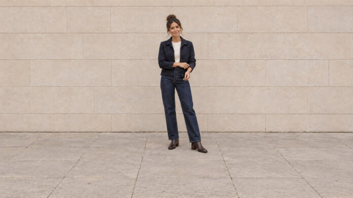 Woman with her hair in a high bun adjusting a navy jacket cuff, wearing straight-leg jeans and brown ankle boots.