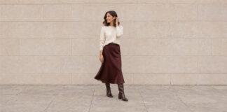 Woman turning in an ivory jumper and burgundy satin midi skirt with brown knee-high boots, lifting a hand to her hair.