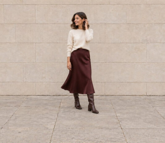 Woman turning in an ivory jumper and burgundy satin midi skirt with brown knee-high boots, lifting a hand to her hair.