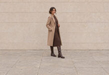 Woman with a low chignon wearing a chocolate knitted dress, camel coat and brown knee-high boots in a three-quarter pose.