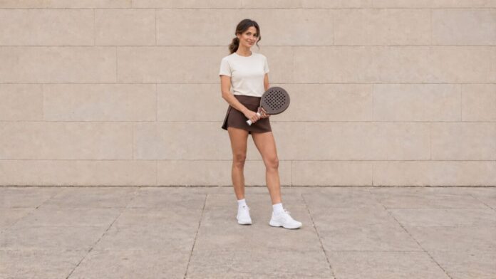 Woman facing the camera in an ivory padel top and chocolate skort, holding a racket in front of a beige stone wall